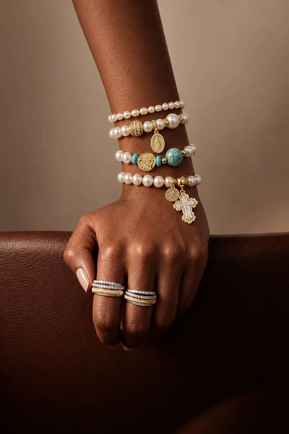 Mão com anéis e pulseiras de pérolas, detalhes dourados, contas azul-turquesa e pingentes religiosos.